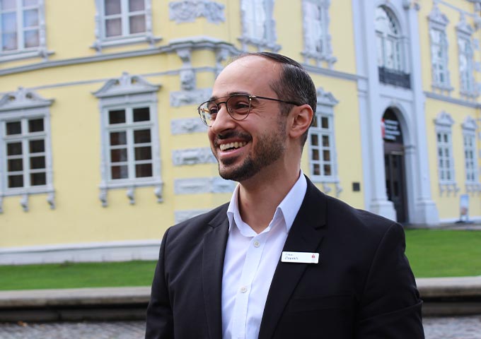 Ein lächelnder Mann im Anzug und mit Brille steht im Freien vor einem gelben historischen Gebäude mit weißen Fenstern und Verzierungen. Er trägt ein Namensschild und scheint den Moment zu genießen.
