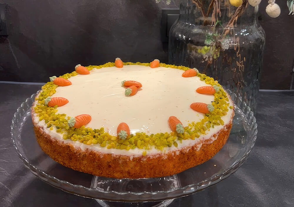 Ein runder Karottenkuchen mit weißem Zuckerguss, verziert mit kleinen Marzipankarotten und gehackten Pistazien am Rand, präsentiert auf einem Tortenständer aus Glas.