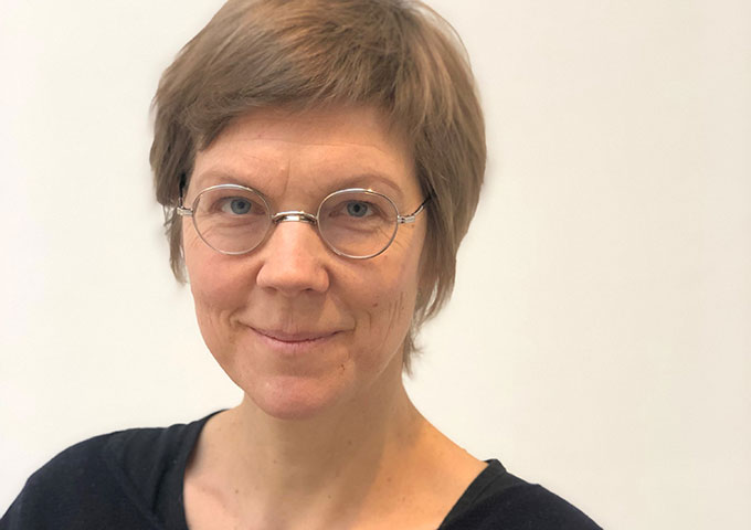 Eine Person mit kurzen hellbraunen Haaren und einer runden Brille, die ein schwarzes Oberteil trägt, lächelt vor einem schlichten hellen Hintergrund sanft in die Kamera.