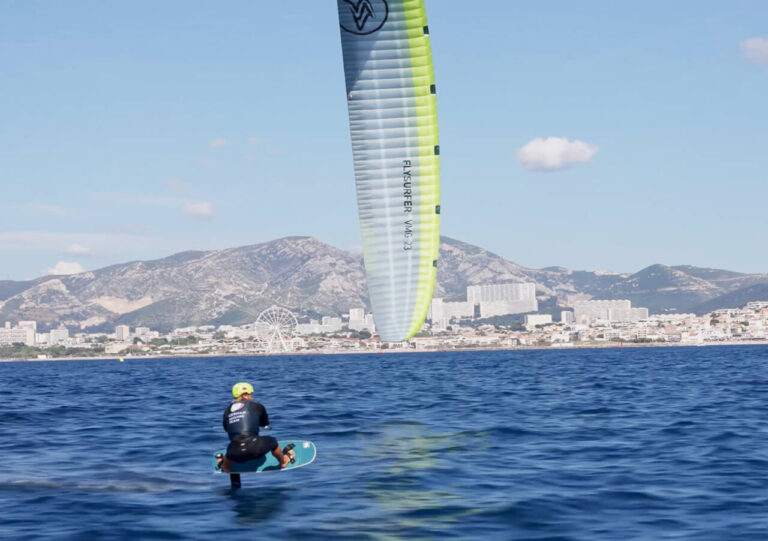 Profi-Kitesurfer Jannis Maus, in Neoprenanzug und Helm, fährt auf einem Tragflächenboot mit einem großen Drachen auf dem Meer. Im Hintergrund sind eine Stadtlandschaft und Berge unter einem klaren blauen Himmel zu sehen, die auf Olympia-Träume hindeuten.