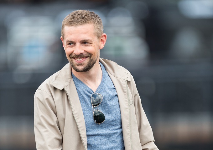 Klaas Heufer-Umlauf, mit kurzen hellbraunen Haaren und einem Bart, lächelt, während er eine helle Jacke, ein blaues T-Shirt und eine Sonnenbrille trägt, die an seinem Hemd hängt. Der Hintergrund ist unscharf.