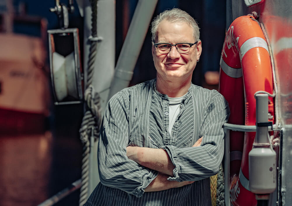 Ein Mann mittleren Alters mit Brille und grauem Haar, der ein gestreiftes Hemd trägt, steht mit verschränkten Armen an einem Rettungsring auf einem Boot während der Oldenburger Land Nacht und lächelt in die Kamera. Der Hintergrund zeigt unscharfe Bootsausrüstung und Wasser.