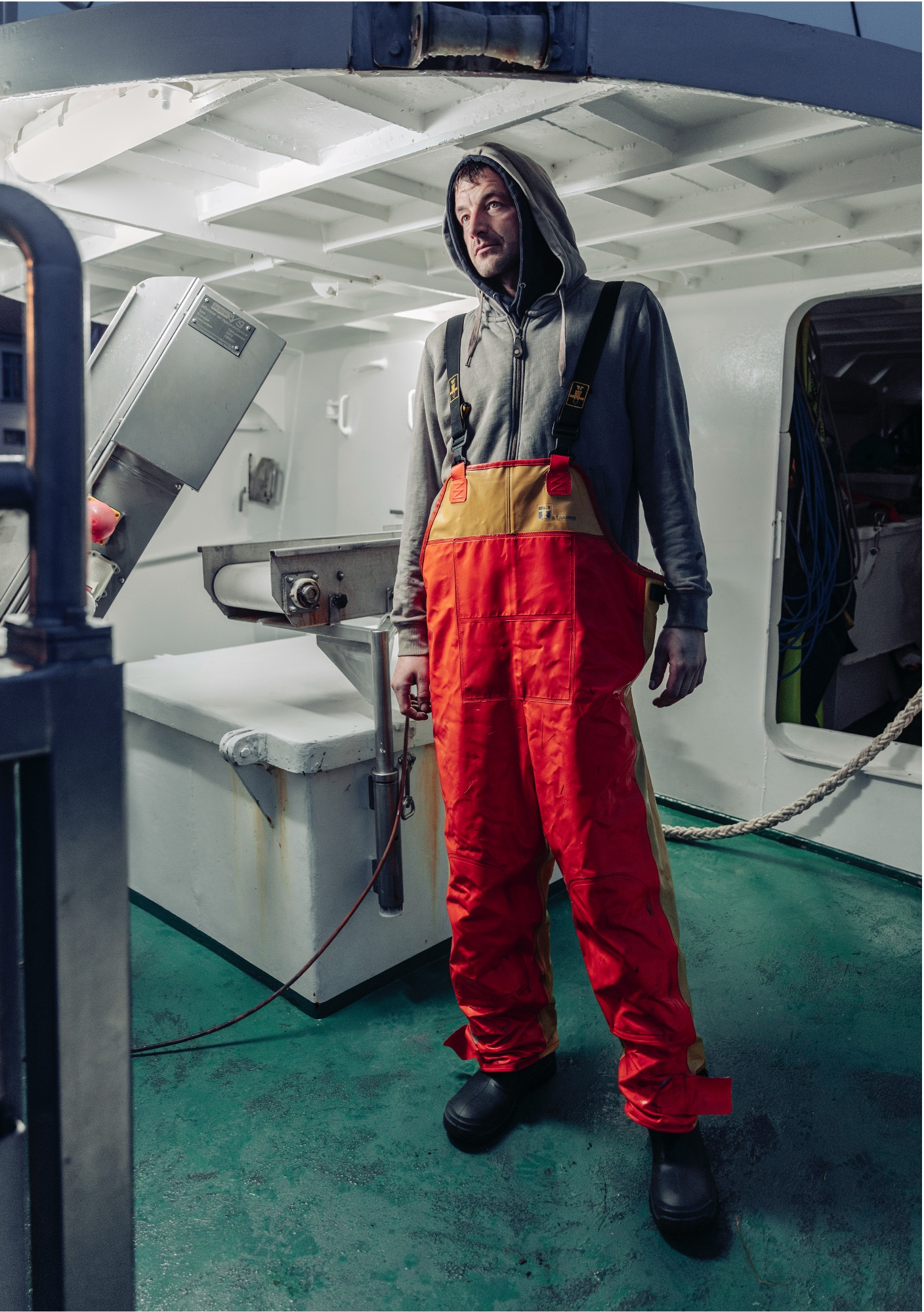 Ein Mann in einem orangefarbenen, wasserdichten Overall und einem grauen Kapuzenpulli steht auf dem Deck eines Bootes, umgeben von Ausrüstung und heller Deckenbeleuchtung.