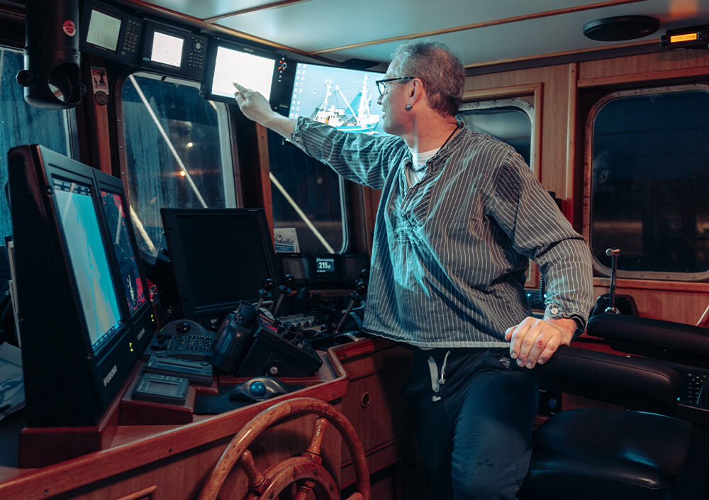 Eine Person sitzt in einem Schiffskontrollraum am Steuer, trägt eine Brille und ein gestreiftes Hemd und streckt ihre Hände in Richtung der mit blauem Licht beleuchteten Navigationsbildschirme und Monitore. Holztafeln und Schiffskontrollen umgeben sie.