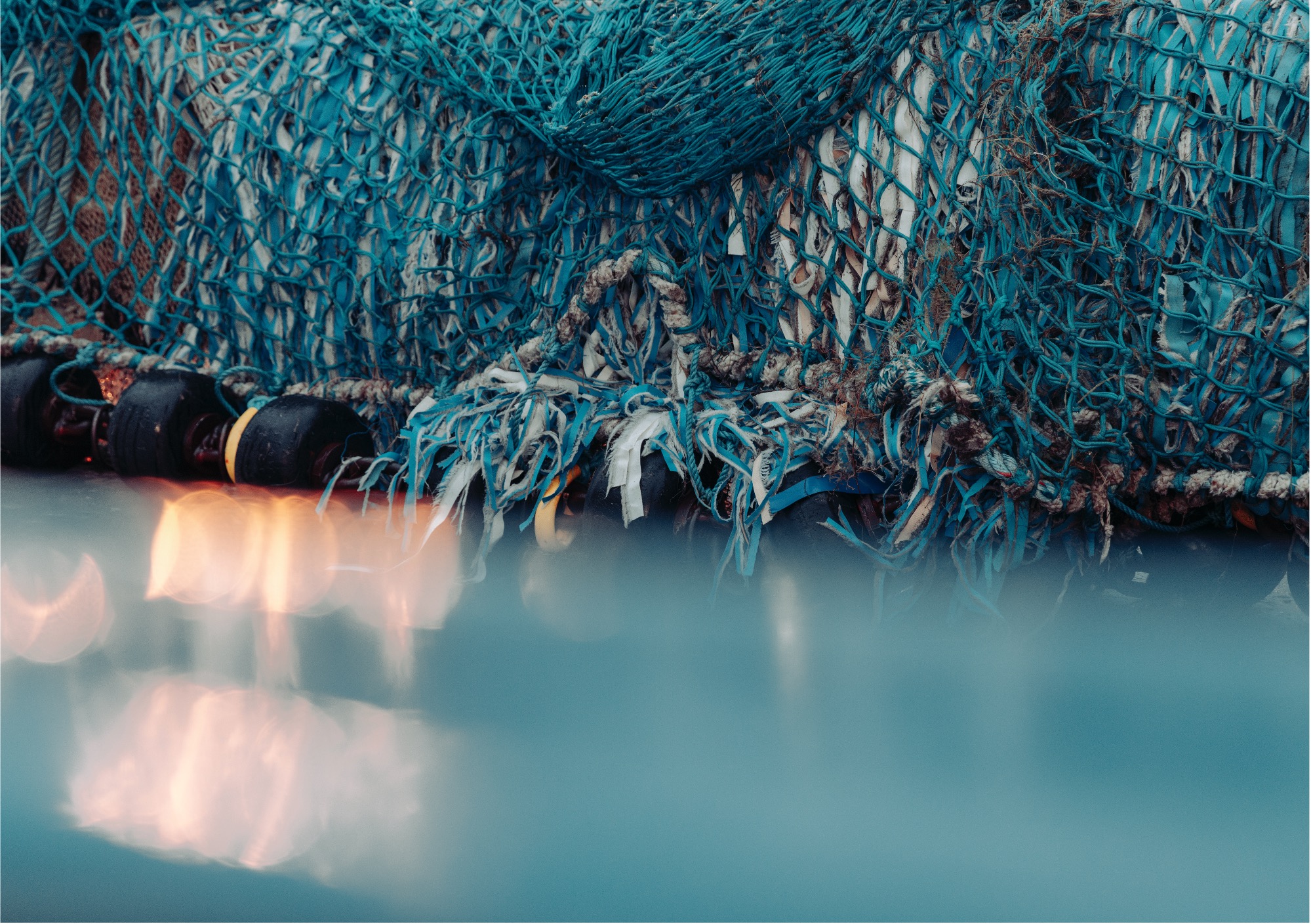 Nahaufnahme eines verhedderten blauen Fischernetzes mit schwarzen Schwimmern, ausgefransten Rändern und einigen Trümmern, das auf einer spiegelnden nassen Oberfläche mit verschwommenen Lichtern im Vordergrund liegt.