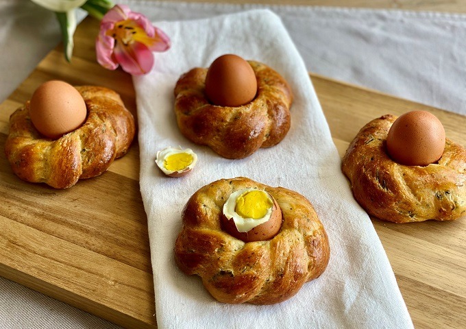 Vier geflochtene Brötchen, auf denen jeweils ein ganzes braunes Ei liegt, werden auf einem weißen Tuch und einem Holzbrett angeordnet. Ein Ei ist aufgeschlagen und gibt den Blick auf ein gelbes Eigelb frei. Eine rosa Tulpe liegt daneben.
