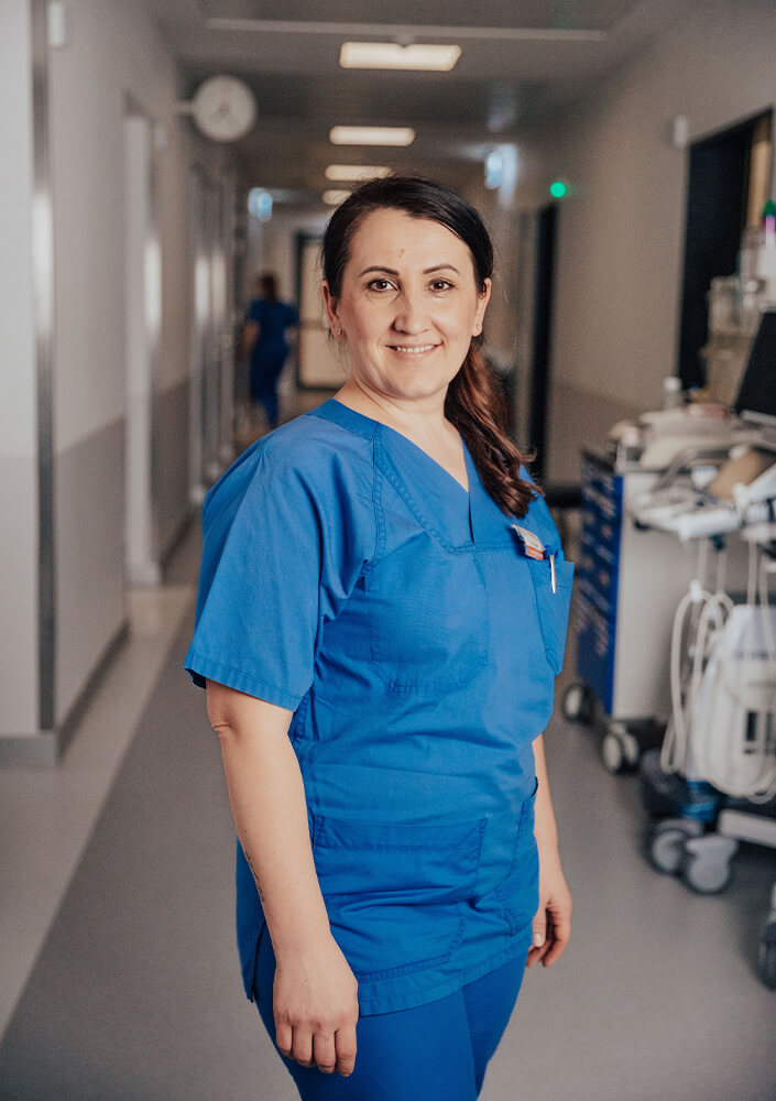 Eine lächelnde Frau in einem blauen Arztkittel steht in einem Krankenhausflur, in dem im Hintergrund medizinische Geräte und helle Deckenleuchten zu sehen sind.
