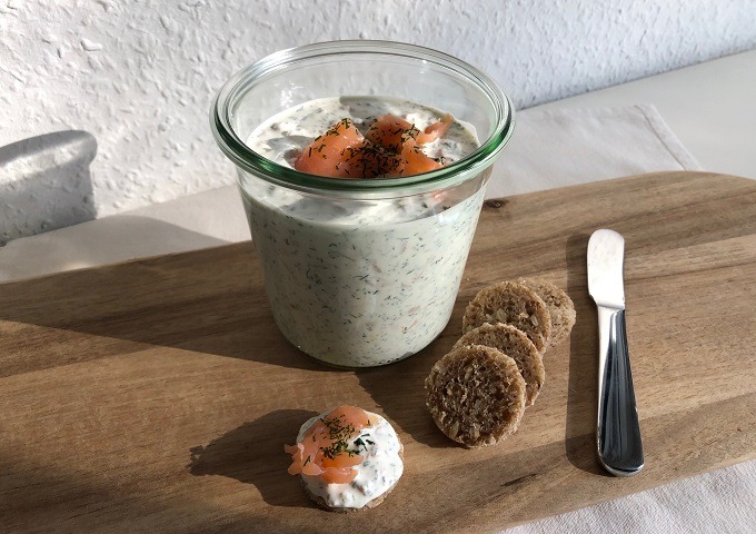 Ein Glas mit einem cremigen Kräuterdip und geräuchertem Lachs steht auf einem Holzbrett, daneben runde Cracker, ein Messer und ein Cracker mit Dip und Lachs. Das Sonnenlicht wirft Schatten auf die weiße Tischplatte.