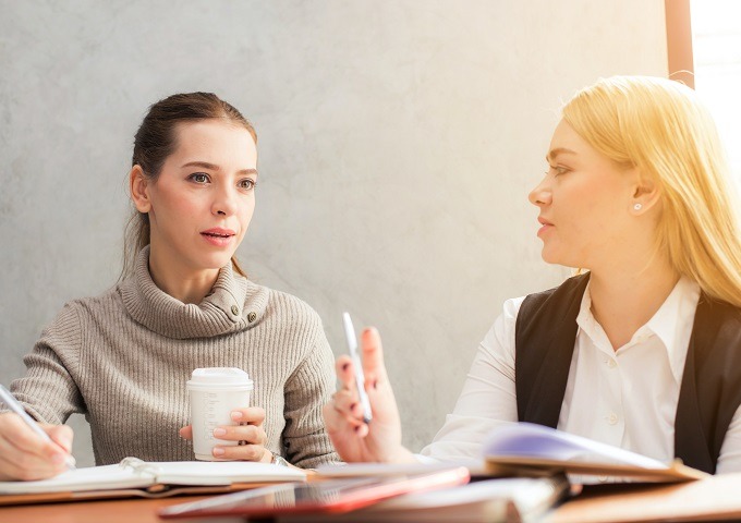 Zwei Frauen sitzen an einem Tisch und führen ein Gespräch. Die eine hält eine Kaffeetasse in der Hand und macht sich Notizen, während die andere mit einem Stift gestikuliert. Beide scheinen in ein Gespräch vertieft zu sein und haben Notizbücher und Papiere vor sich liegen.