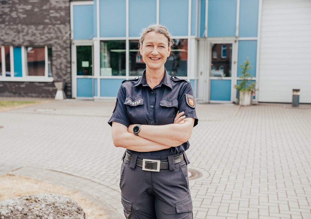 Eine lächelnde Polizeibeamtin mit kurzen grauen Haaren steht vor einem Gebäude, die Arme verschränkt, in einer Marineuniform mit Abzeichen und einer Smartwatch, auf einer gepflasterten Fläche mit Fenstern und einem kleinen Baum hinter ihr.