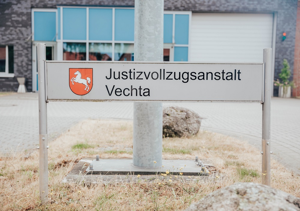 Ein Schild vor einem Gebäude mit der Aufschrift Justizvollzugsanstalt Vechta und einem roten Schild mit einem weißen Pferd weist auf eine Justizvollzugsanstalt in Vechta, Deutschland, hin. Das Gelände ist gepflastert, mit etwas Gras und Steinen.