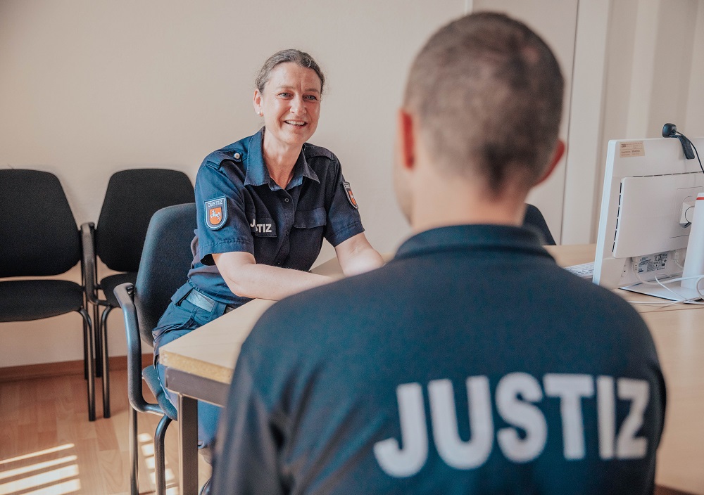 Eine Frau in dunkler Uniform mit Dienstmarke lächelt, während sie in einem Büro mit leeren Stühlen und einem Computer an einem Schreibtisch sitzt, gegenüber einer anderen Person in einem JUSTIZ-Hemd.