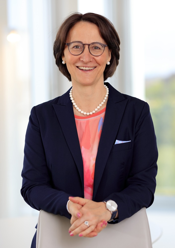 Eine Frau mit kurzen braunen Haaren und Brille, die einen marineblauen Blazer, eine rosa gemusterte Bluse, eine Perlenkette und eine Uhr trägt, lächelt, während sie in einem Haus mit natürlichem Licht im Hintergrund steht.