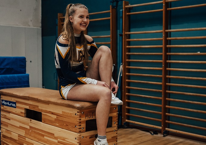 Eine lächelnde Cheerleaderin in Uniform sitzt auf einem hölzernen Sprungturm und bindet ihren Schuh in einer Turnhalle mit Sprossenwand im Hintergrund.