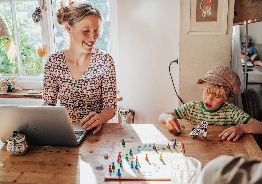 Eine Frau arbeitet an einem Laptop an einem Holztisch und lächelt einem kleinen Jungen zu, der mit einem Spielzeugflugzeug und einem Brettspiel spielt. Das Sonnenlicht fällt durch ein nahe gelegenes Fenster und schafft eine gemütliche, heimelige Atmosphäre.