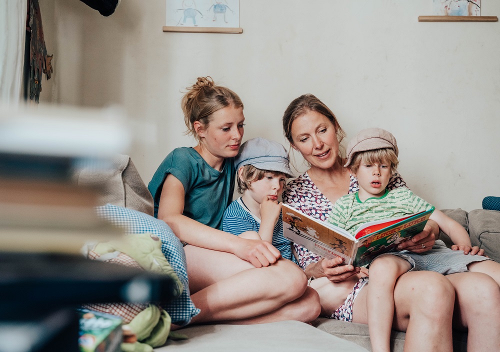 Eine Frau sitzt auf einer Couch und liest zwei Jungen mit Hüten ein Kinderbuch vor, während ein Mädchen daneben sitzt, alle wirken beschäftigt und gemütlich.