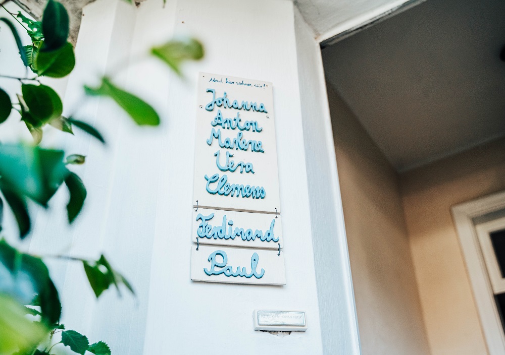 Eine weiße Wand mit handgeschriebenen Namensschildern, auf denen Johanna, Anton, Malia, Uta, Clemens, Ferdinand und Paul in blauer Schrift stehen, neben einer Tür und einigen grünen Pflanzenzweigen.