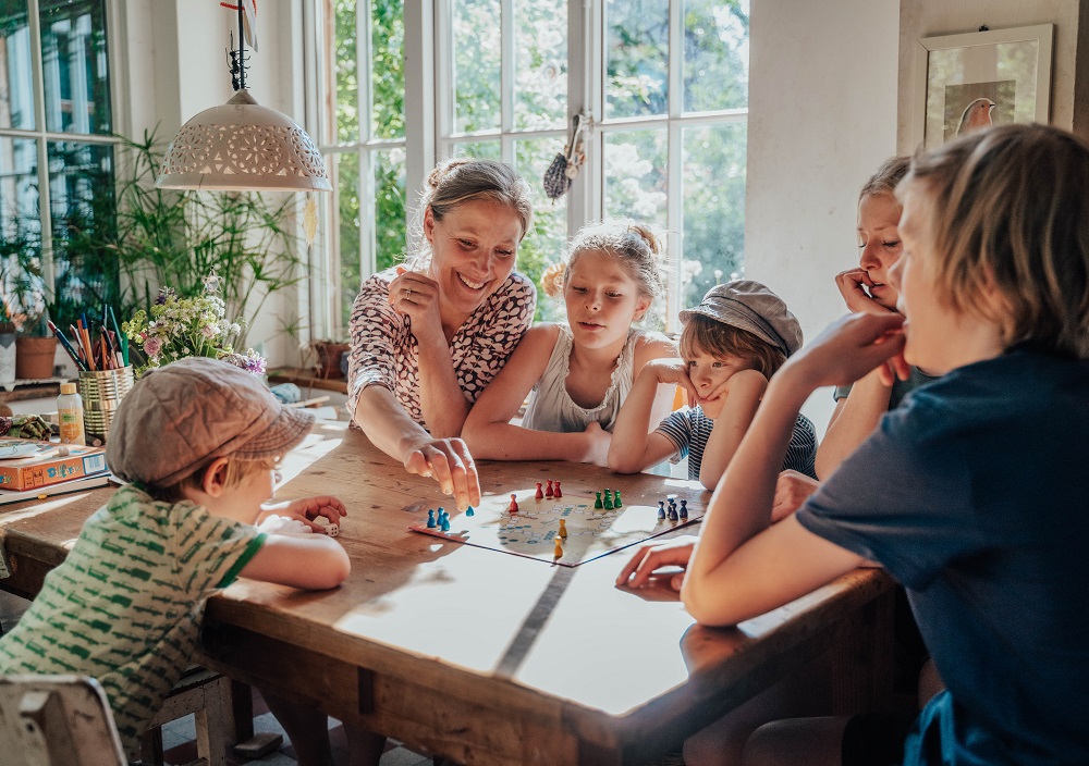 Eine Frau und fünf Kinder versammeln sich um einen Holztisch in einem sonnenbeschienenen Raum und spielen gemeinsam ein buntes Brettspiel. Alle scheinen konzentriert und engagiert zu sein und das Spiel in einem gemütlichen, mit Pflanzen gefüllten Raum zu genießen.