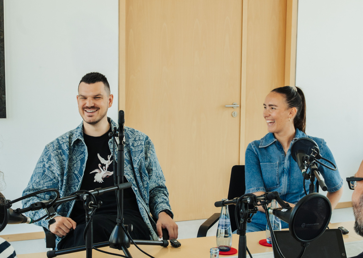 Zwei Personen sitzen mit Mikrofonen, Kopfhörern und Wasserflaschen an einem Tisch, lächeln und unterhalten sich, als würden sie in einem hellen, modernen Raum einen Podcast oder ein Interview aufnehmen.
