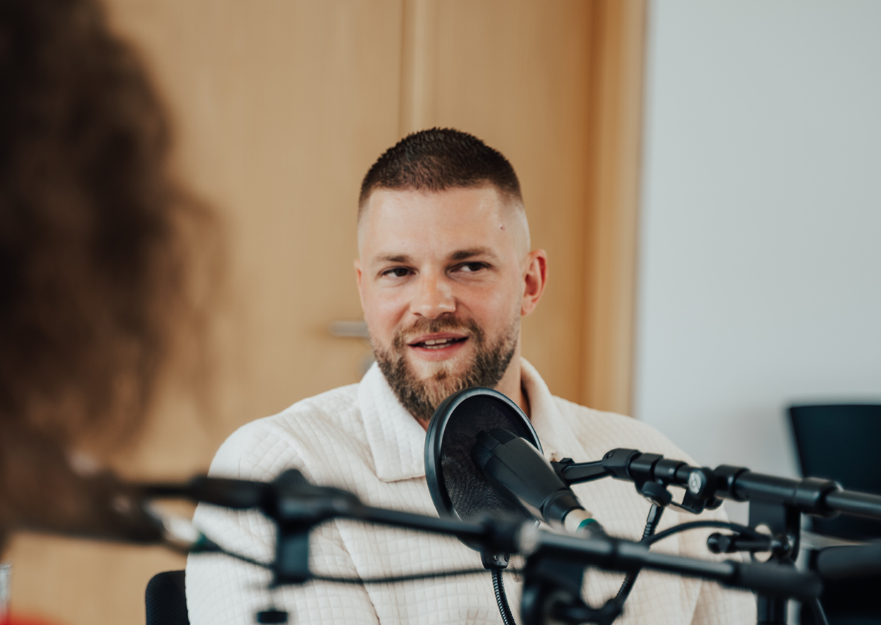 Ein Mann mit gestutztem Bart und kurzen Haaren spricht in ein Mikrofon während einer Podcast- oder Radioaufnahme. Im Vordergrund ist eine weitere Person zu sehen, die unscharf ist. Die Umgebung scheint ein Studio oder ein Büro zu sein.
