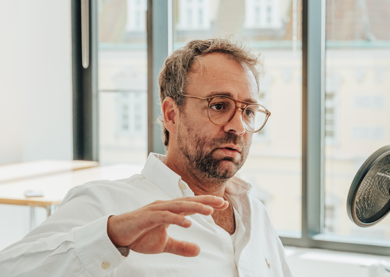 Ein Mann mit hellbraunem Haar und Brille, der ein weißes Hemd trägt, gestikuliert mit seiner Hand, während er in einem hellen Büro mit großen Fenstern im Hintergrund spricht.