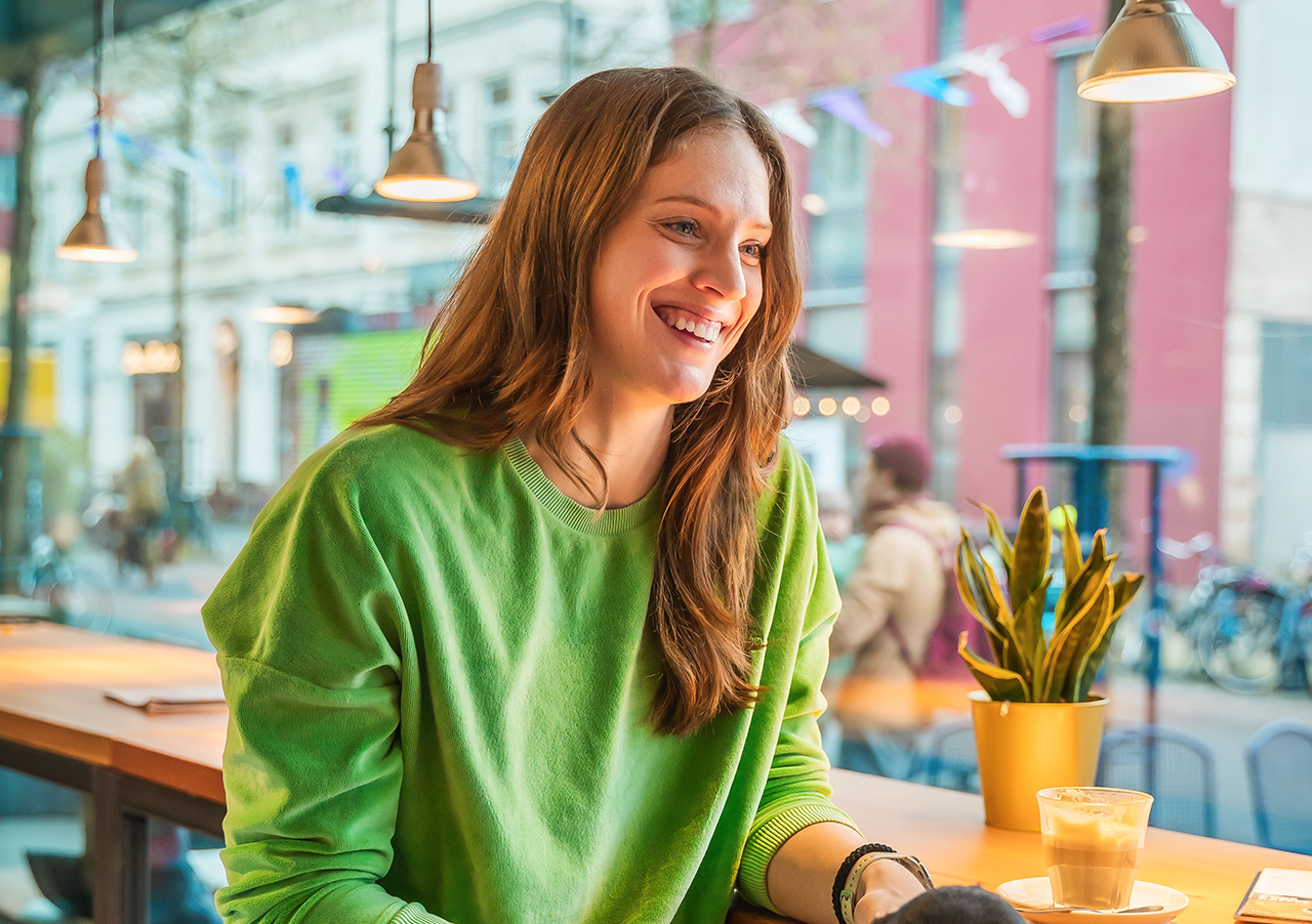 Eine junge Frau in einem hellgrünen Sweatshirt lächelt, während sie mit einem Kaffee und einer Topfpflanze am Tresen eines Cafés sitzt, dessen große Fenster eine Straße in der Stadt zeigen.