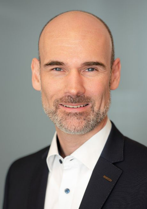 Ein Mann mit Glatze und kurzem Bart lächelt, trägt ein weißes Hemd und eine dunkle Anzugsjacke und posiert vor einem schlichten grauen Hintergrund.