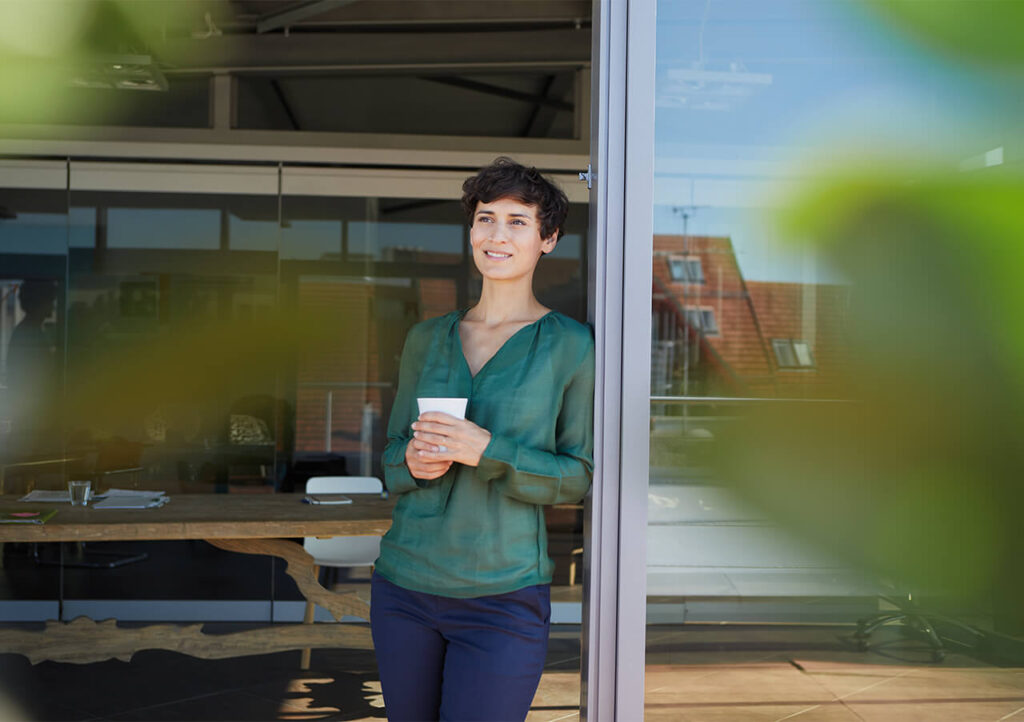 Eine Person mit kurzen dunklen Haaren, die eine grüne Bluse und eine dunkle Hose trägt, steht an einer offenen Glastür, hält eine Tasse und lächelt. Der Hintergrund zeigt ein modernes Büro mit einem Holztisch und großen Fenstern.