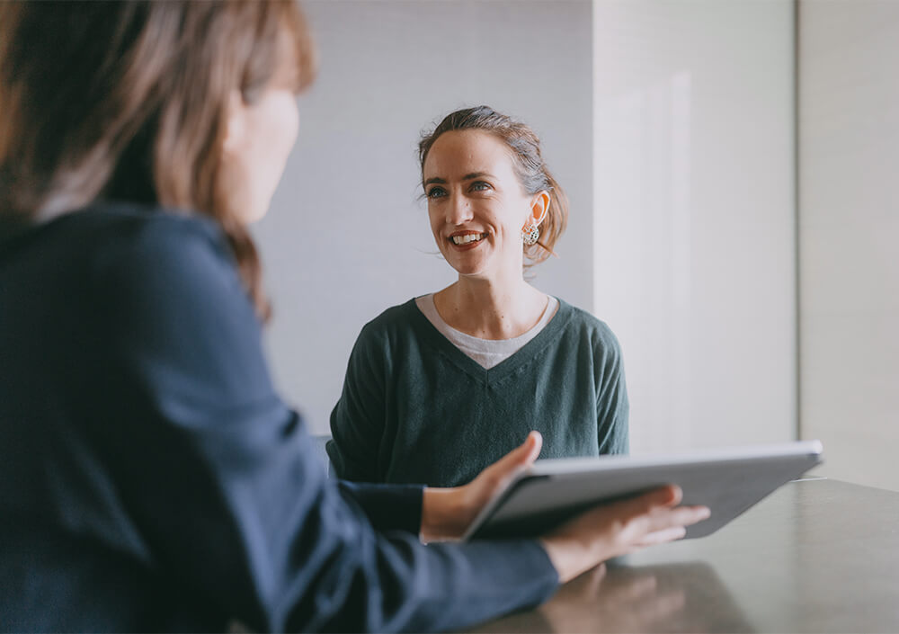 Zwei Frauen sitzen an einem Tisch und führen ein Gespräch. Die eine lächelt freundlich, während die andere, deren Gesicht unscharf ist, ein Tablet hält. Die Umgebung wirkt hell und professionell, möglicherweise während einer Besprechung oder eines Vorstellungsgesprächs.