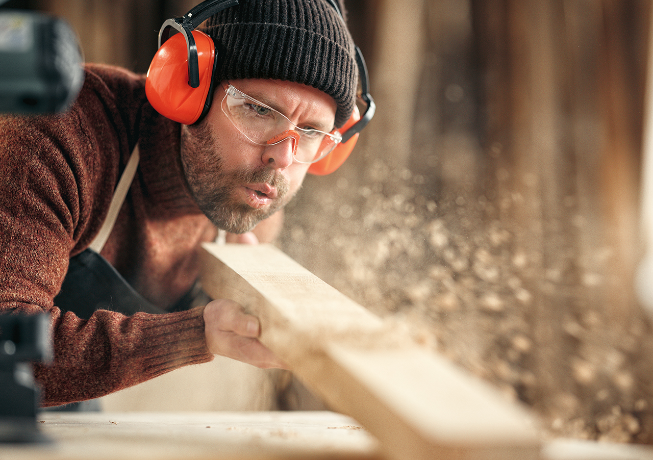Ein Mann, der eine Schutzbrille, eine Mütze und orangefarbene Ohrenschützer trägt, bläst bei der Arbeit in einer Werkstatt Sägespäne von einem Holzbrett. Um ihn herum fliegen Holzspäne in die Luft.