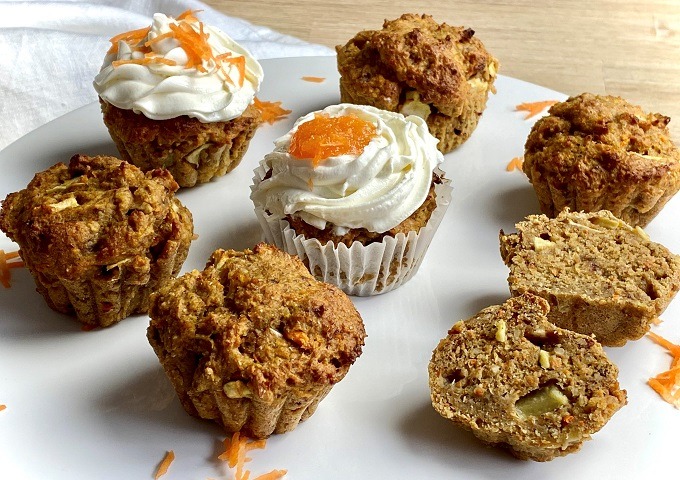 Ein Teller mit Karottenmuffins, einige mit weißem Zuckerguss und Karottenraspeln obenauf, andere ohne. Ein Muffin ist halbiert und zeigt das feuchte, strukturierte Innere mit Karottenstücken und Nüssen.