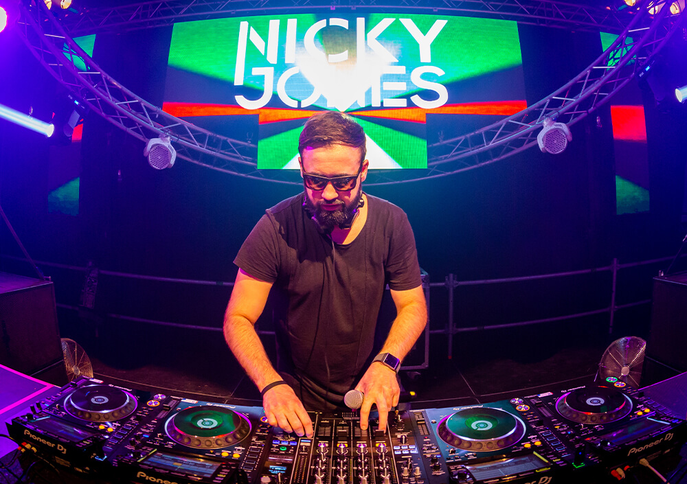 Ein DJ im Club, der eine Brille und Kopfhörer trägt, steht auf der Bühne und mischt Musik an einem DJ-Pult. Helle Lichter und ein bunter Bildschirm hinter ihm zeigen den Namen Nicky Jones an.