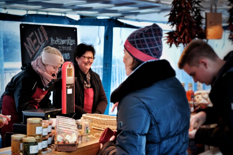 Zwei Frauen hinter einem Marktstand lächeln, während sie bei der Oldenburger Land Nacht Kunden bedienen. Vorne stehen Gläser mit Waren, im Hintergrund ein Kreideschild, während auf der anderen Seite der Theke zwei Kunden stehen.