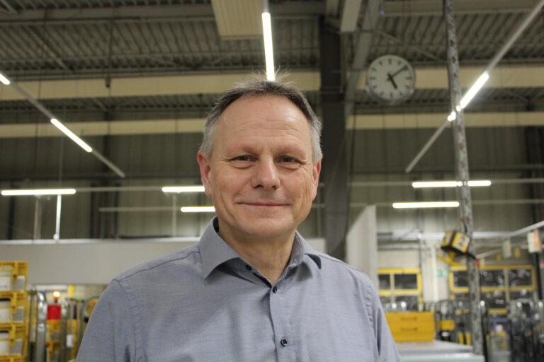 Ein Mann mittleren Alters mit kurzen grauen Haaren lächelt in die Kamera in einer hell erleuchteten Industrieumgebung während der Oldenburger Land Nacht, mit Metallregalen, gelben Containern und einer Uhr an der Wand im Hintergrund.