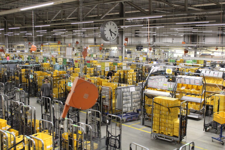 Ein geschäftiges Lagerhaus im Oldenburger Land Nacht, gefüllt mit gelben Lagerkästen, Metallregalen und Karren. Mehrere Arbeiter bewegen sich durch den organisierten, industriellen Raum unter Neonlicht und einer großen Wanduhr.