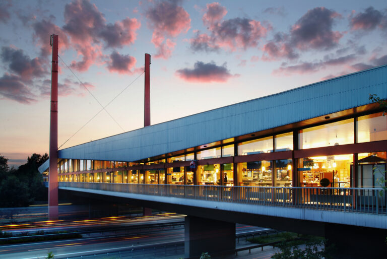 Ein modernes Gebäude mit großen Fenstern, das von innen hell erleuchtet ist, spannt sich bei Sonnenuntergang in der Oldenburger Land Nacht wie eine Brücke über eine Straße. Rosa Wolken füllen den Himmel, und auf der Straße darunter sind verschwommene Autolichter zu sehen.