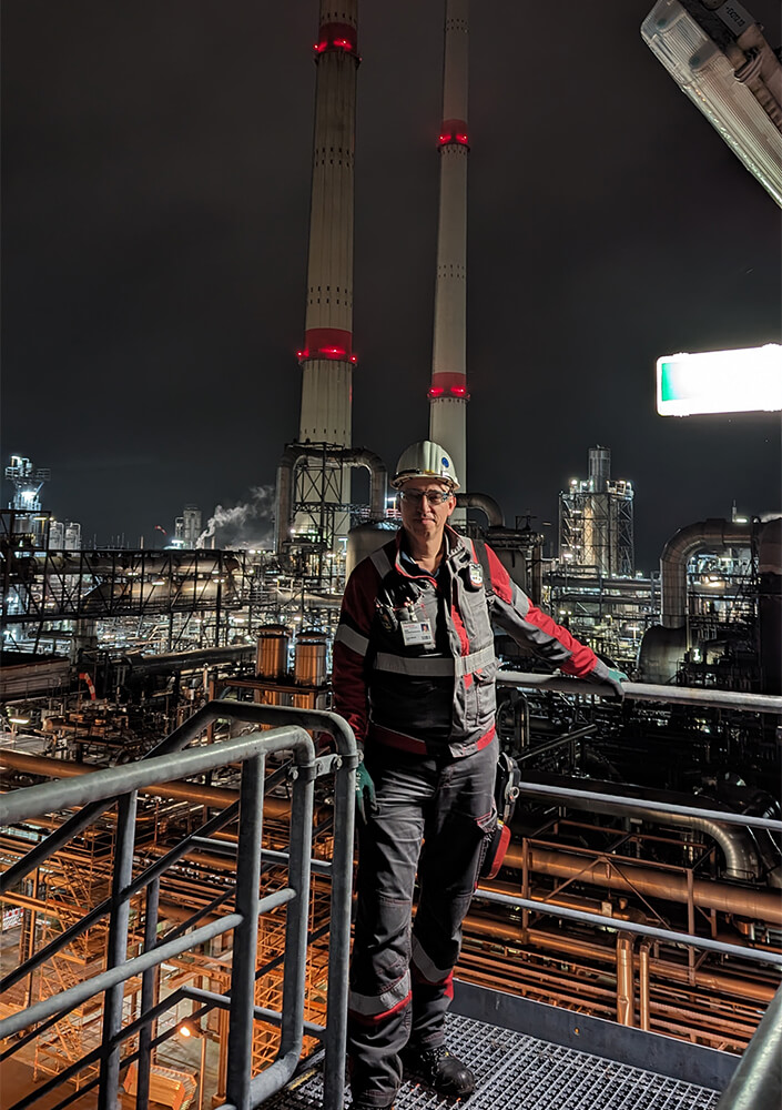 Ein Arbeiter in Schutzkleidung und mit Helm steht auf einer Metallplattform in einer nächtlichen Industrieanlage im Oldenburger Land. Große Schornsteine und beleuchtete Raffineriegebäude leuchten gegen den Nachthimmel.