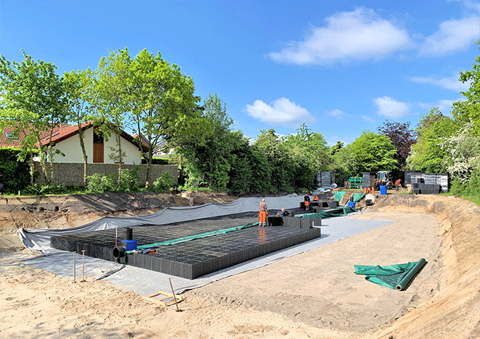 Baustelle mit Arbeitern, die modulare Kunststoffkisten in einer großen rechteckigen Grube zusammensetzen, umgeben von Bäumen, Häusern und Baumaterialien an einem sonnigen Tag.