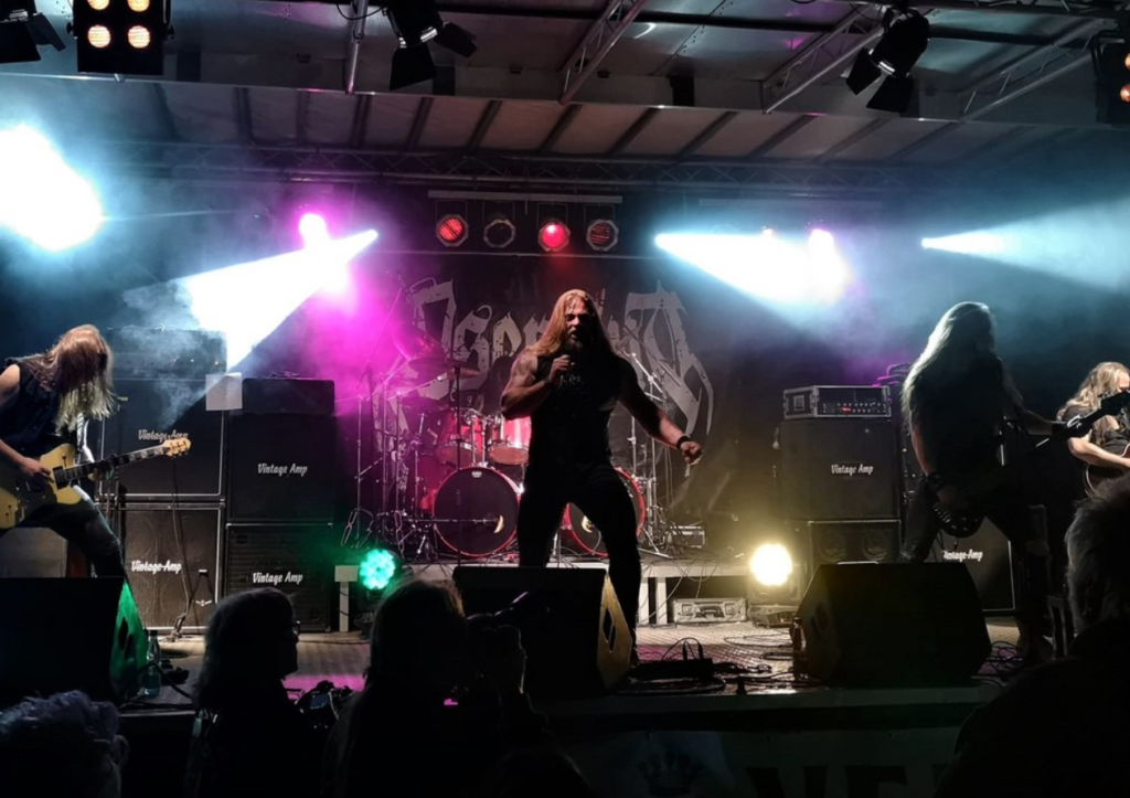 Eine Heavy-Metal-Band spielt energisch auf einer mit Rauch gefüllten Bühne mit bunten Lichtern, Verstärkern und einem Hintergrund mit ihrem Logo, während das Publikum im Vordergrund zusieht.