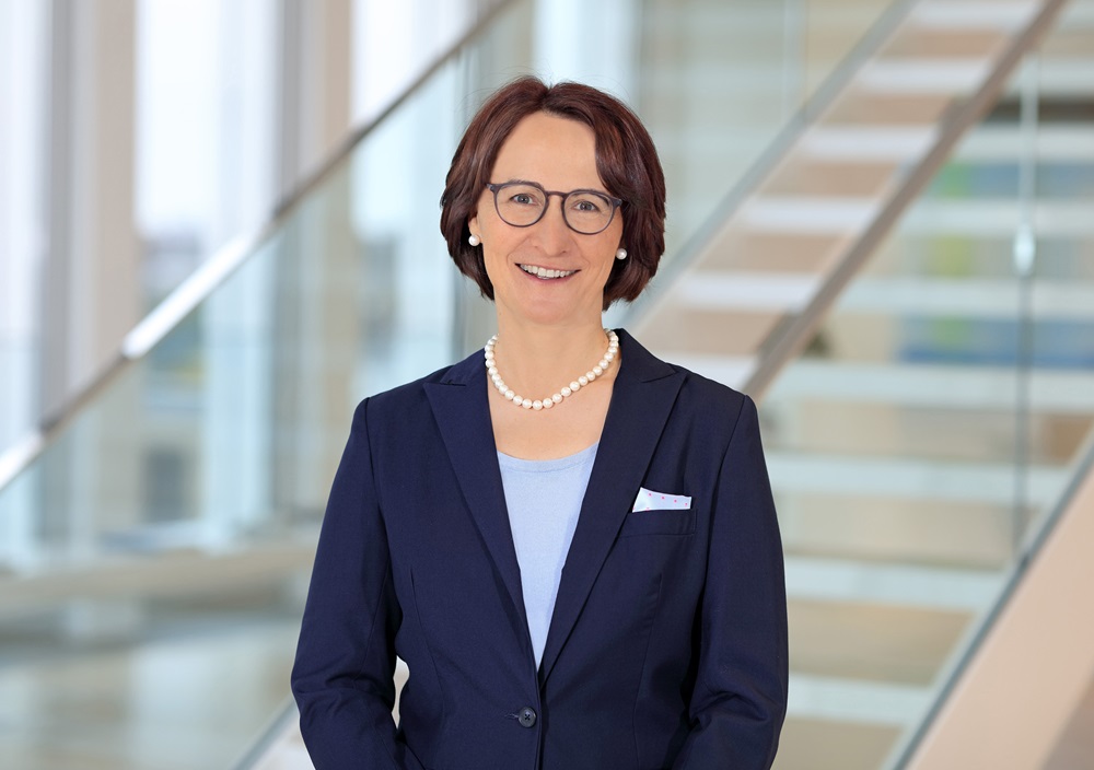 Eine Frau mit kurzen braunen Haaren und Brille, die einen marineblauen Blazer, ein hellblaues Top und eine Perlenkette trägt, lächelt, während sie vor einer modernen Treppe mit Glasgeländer steht.