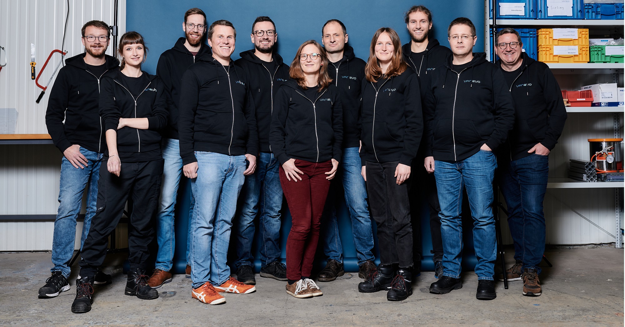 Eine Gruppe von elf Mitarbeitern der Firma Vanevo in schwarzen Kapuzenpullis und legerer Kleidung steht in einer Halle und posiert lächelnd für ein Teamfoto vor einer blau-weißen Wand, an der im Hintergrund Regale und Werkstattwerkzeuge zu sehen sind.