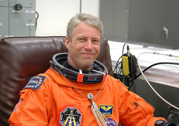 Ein lächelnder Astronaut sitzt auf einem Stuhl und trägt einen orangefarbenen Raumanzug mit Missionsaufnähern und einem teilweise geöffneten Helmkragen in einem Vorbereitungsraum.