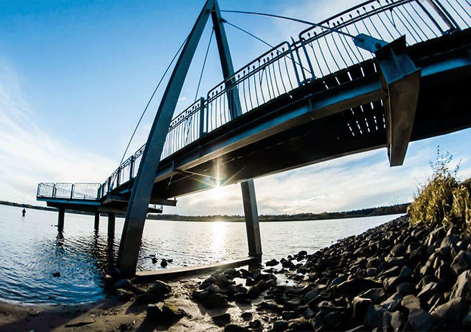 Eine moderne Fußgängerbrücke mit Metallgeländer überquert einen Fluss bei Sonnenuntergang. Steine säumen das Ufer im Vordergrund, und die Sonne scheint durch die Brückenkonstruktion und wirft Spiegelungen auf das Wasser.