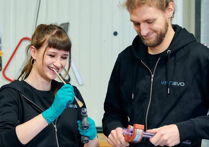 Zwei Personen in schwarzen Kapuzenpullovern arbeiten zusammen in einem VANEVO-Labor. Die Frau lächelt, während sie eine Bürste und ein Röhrchen in der Hand hält, und trägt blaugrüne Handschuhe. Der Mann benutzt eine Klemme an einem Rohr. Beide scheinen konzentriert und engagiert bei der Arbeit zu sein.