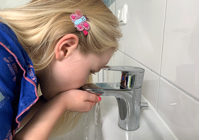 Ein junges Mädchen mit blondem Haar und einer rosafarbenen Haarspange trinkt Wasser aus einem Wasserhahn im Badezimmer, wobei sie das Wasser mit den Händen schöpft, während es aus dem Hahn läuft.
