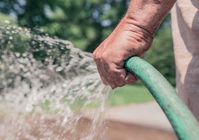 Nahaufnahme einer Person, die einen grünen Gartenschlauch hält, der an einem sonnigen Tag im Freien Wasser versprüht, wobei Bäume und Gras im Hintergrund verschwimmen.