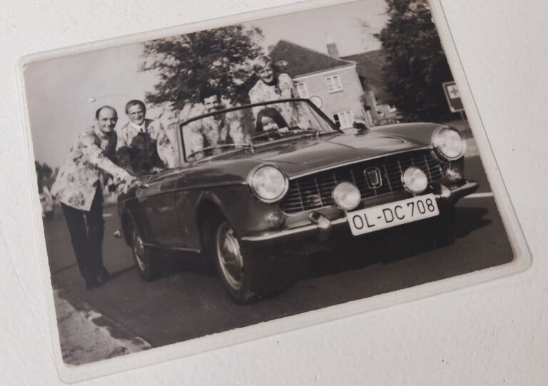 Ein Schwarz-Weiß-Foto zeigt vier lächelnde Menschen um einen Oldtimer-Cabrio mit dem Kennzeichen OL-DC 708 - einen der Wendepunkte im Leben - auf einer von Häusern und Bäumen gesäumten Straße im Hintergrund.