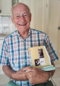 Ein älterer Mann mit kurzen weißen Haaren lächelt und hält eine Mappe mit Fotos und Papieren in der Hand - vielleicht denkt er über Wendepunkte im Leben nach. Er trägt ein bunt kariertes Hemd und sitzt in einem hellen Raum mit hellen Wänden.