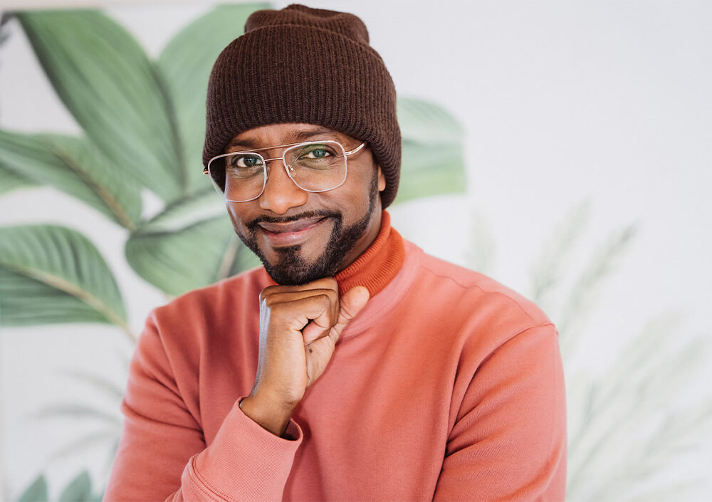 Ein lächelnder Mann mit Brille, brauner Strickmütze und rosa Sweatshirt stützt sein Kinn auf seine Hand. Große grüne Blätter verschwimmen im Hintergrund und tragen zu dieser gemütlichen Patchwork-Heimat-Atmosphäre bei.
