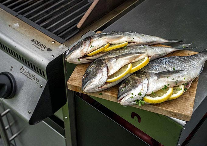 Drei ganze rohe Fische, gefüllt mit Zitronenscheiben und Kräutern, liegen auf einem Holzbrett neben einem Metallgrill und sind bereit zum Garen.